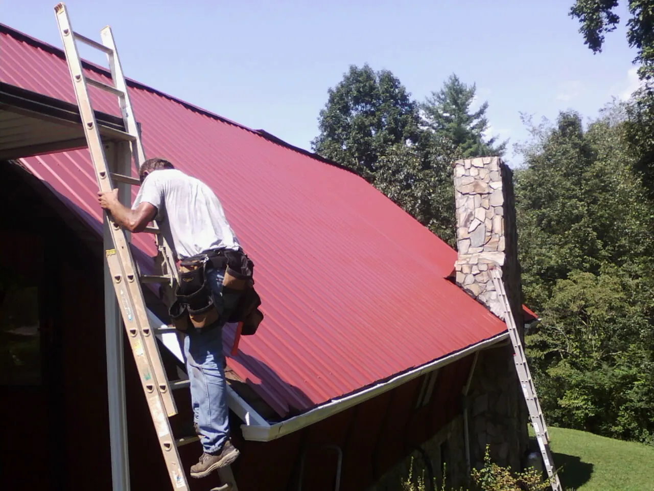 Roofer on a ladder beside a red metal roof and stone chimney — Soffit & Fascia Repair by Roofing Brothers in Cut Off